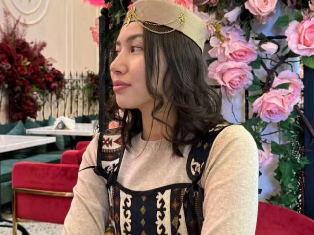 StephanyMays, avec ses cheveux bruns ondulés, portant une pièce de tête dorée, une chemise beige et une robe noire à motifs, regardant sur le côté, dans une pièce avec des roses roses et des meubles rouges..
