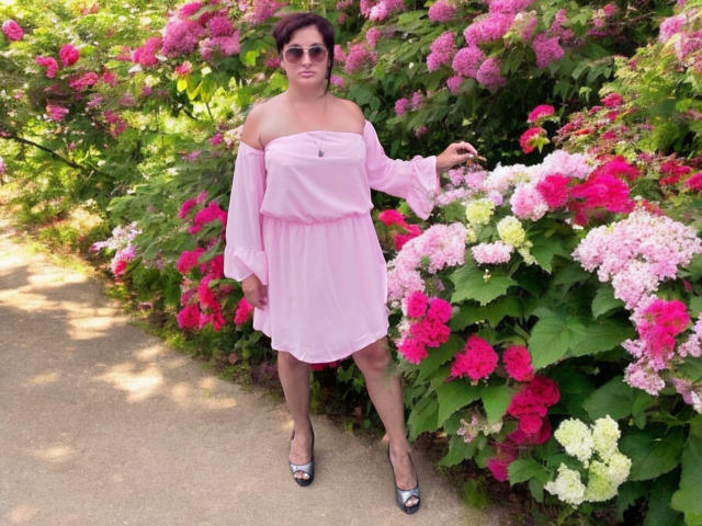 KelliRedhaer stands in a garden wearing a pink off-shoulder dress with puffed sleeves, holding a pink hydrangea. She has short brown hair, sunglasses, and silver peep-toe heels.