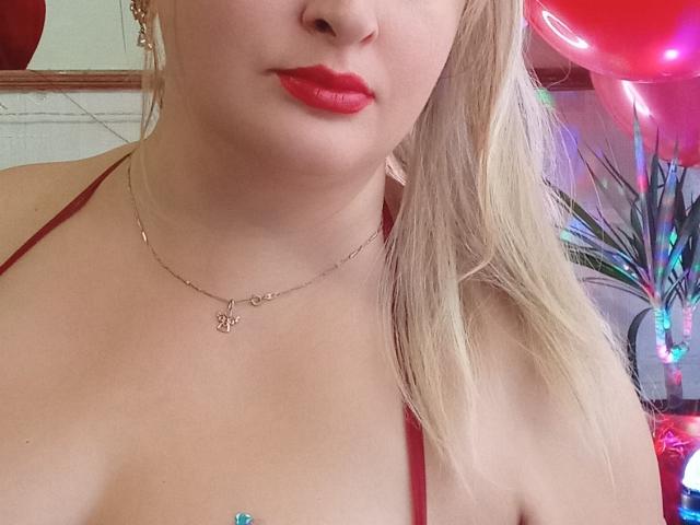 El rostro de KelliRedhaer, con lápiz labial rojo, cabello rubio y un top halter rojo, con un collar dorado y escote visible, sobre un fondo colorido con luces..