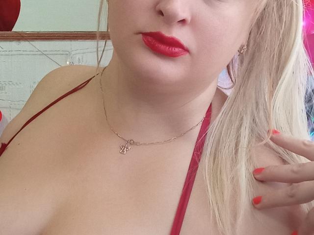 KelliRedhaer con rossetto rosso, capelli biondi, reggiseno rosso, collana dorata e smalto rosso, che mostra i suoi seni e la sua mano. Ha un viso carino e un leggero sorriso..