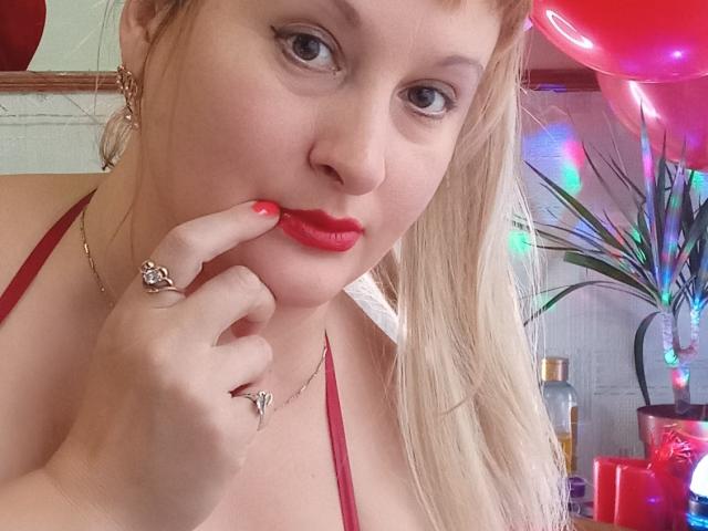 KelliRedhaer, blonde with red lipstick, touches her red lips with a finger, showing off a ring and necklace, wearing a red halter top, with colorful lights in the background.