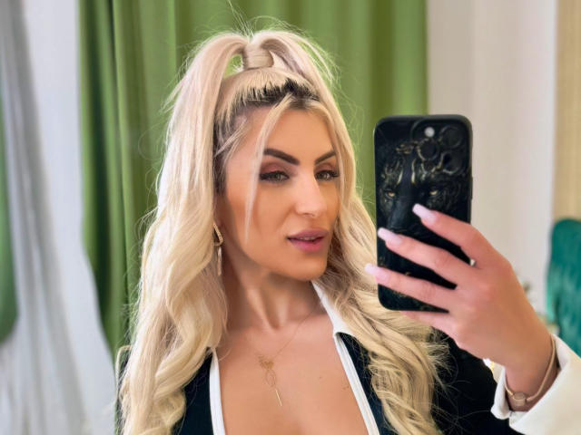 JessycaPearl con lunghi capelli biondi raccolti in una coda alta, che tiene in mano un telefono nero, indossa una maglietta nera e bianca, mostrando il seno, con tende verdi sullo sfondo..
