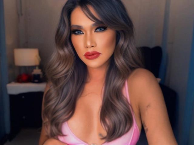 HornyCumTs with long, wavy gray-brown hair, blue eyes, and red lips, wearing a pink bra, showing her boobs. Her makeup is bold, and she's looking at the camera.