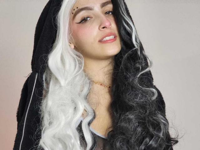 KloehWow a les cheveux noirs et blancs, bouclés, porte un sweat à capuche noir et a un anneau de nez mignon. Elle sourit, avec un eye-liner ailé et un collier en or. Ses seins sont partiellement visibles..
