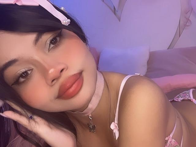 LiliMays with pink lingerie, choker, and headband, has her tits exposed, dark hair, red lips, and a cute smile, lying on a bed, looking at the camera.