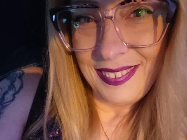 PurplesLips has blonde hair, green eyes, and wears big, clear glasses with purple lipstick. She's smiling with a tattoo on her left arm. Boobs not visible.