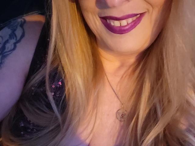 PurplesLips with blonde hair, purple lipstick, black sequined top, necklace, and tattoo on right shoulder, smiling at camera. Boobs visible in top. Face partially cut off.