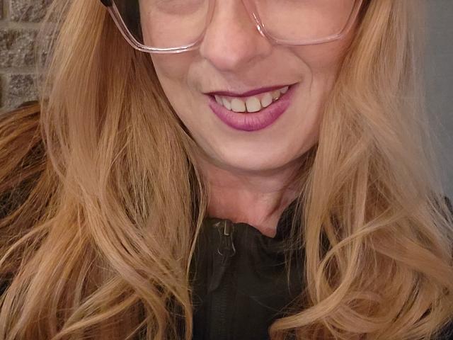 PurplesLips has blonde hair, pink glasses, and purple lipstick, smiling with visible teeth. She's wearing a black shirt, and a brick wall is in the background.