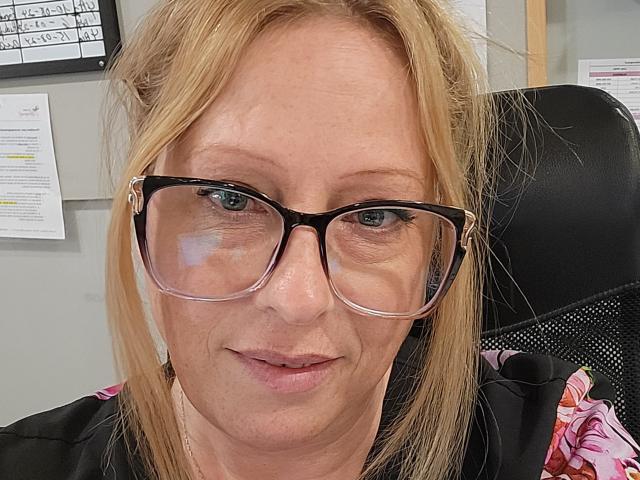 PurplesLips, middle-aged with blonde hair, wearing black glasses and a black office chair, has light skin and blue eyes, with a subtle smile. Boobs not visible, pussy not visible.