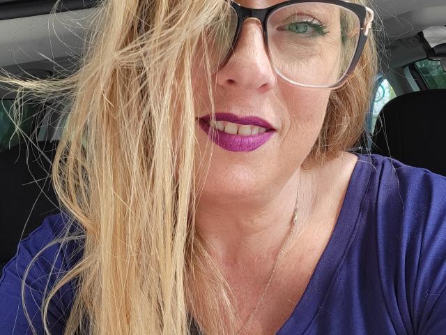 PurplesLips with long blonde hair, black glasses, purple lipstick, and blue shirt, sitting in a car, smiling at the camera, showing green eyes.