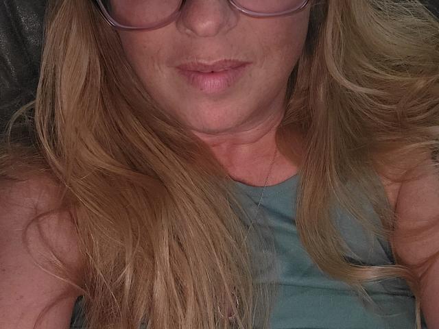 PurplesLips has blonde hair, wears glasses, and a teal tank top, with pouty pink lips, showing a relaxed, slightly sleepy expression. Her tits are not visible.