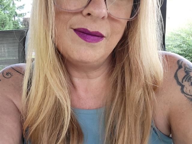 PurplesLips has blonde hair, purple lipstick, glasses, tattoos on both shoulders, and a light blue top, showing a smiling face with a slight double chin.