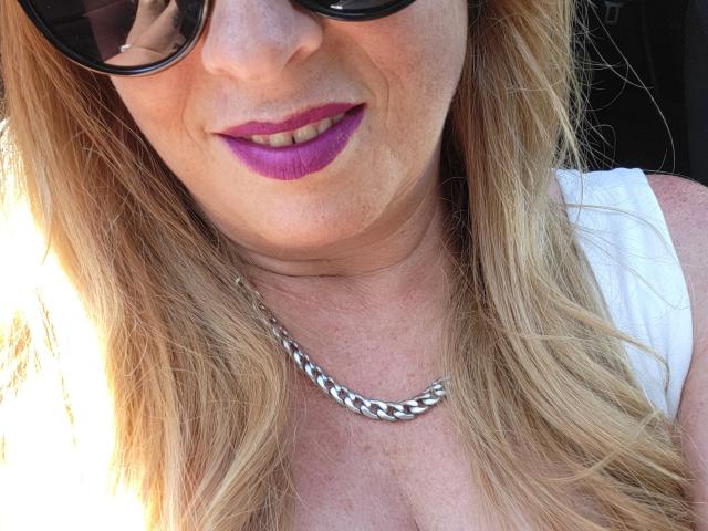 PurplesLips has blonde hair, wearing black sunglasses, white top, and purple lipstick. She's sporting a silver chain necklace, showing a slight smile with sunlit skin.