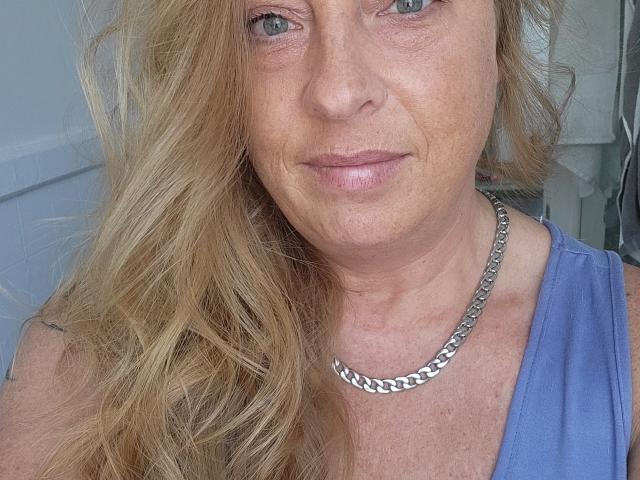 PurplesLips has light blue eyes, wavy blonde hair, and a slight smile. She wears a silver chain necklace and a blue top, showing freckles on her face.