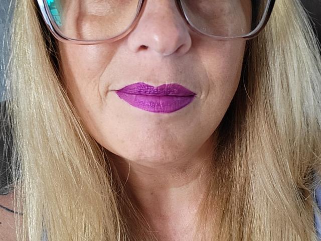 PurplesLips with purple lipstick, wearing glasses, blonde hair, fair skin, slight smile, visible wrinkles, close-up shot, showing nose and mouth, looking down.