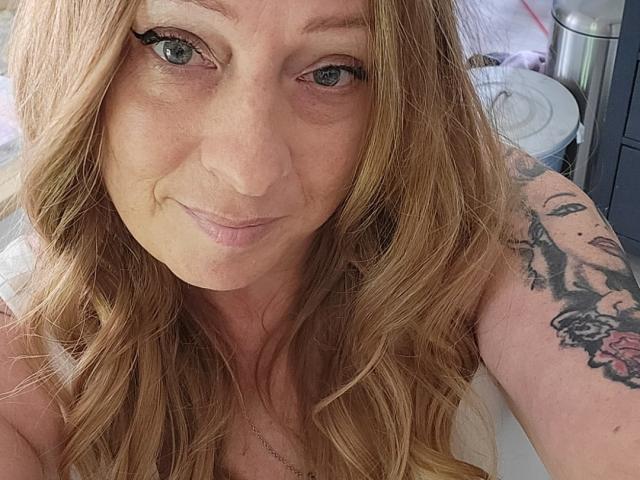 PurplesLips has wavy blonde hair, green eyes, a slight smile, and a tattoo on her right arm. She's wearing a white top and a silver necklace.