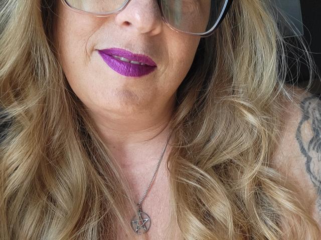 PurplesLips has wavy blonde hair, purple lipstick, glasses, and a silver pentacle necklace, with a tattoo on their right shoulder. Their face is the focus.