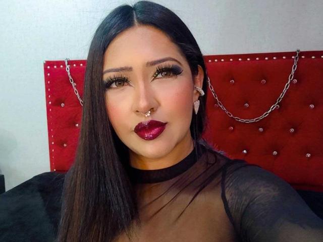 Melanie Morel ha lunghi capelli scuri, occhi marroni, rossetto rosso, una maglia nera trasparente, un piercing al naso e orecchini, ed è seduta contro una testiera rossa con catene e decorazioni. Il seno è visibile..