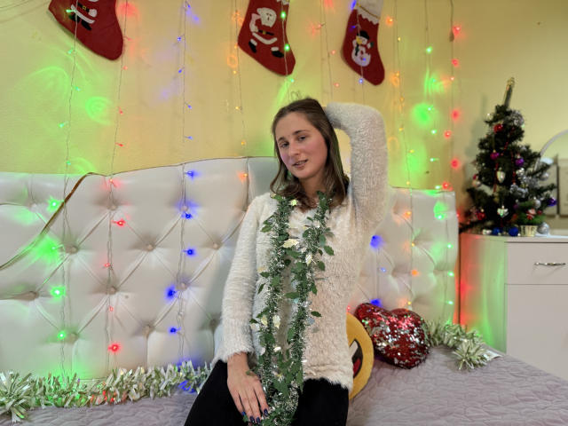 MichelleHotX69 with a cute face, wearing a white fuzzy sweater, sitting on a bed with colorful Christmas lights, green garland, and black pants, looking relaxed.