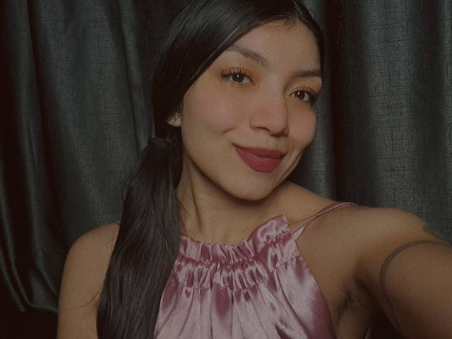 IbbyeMoreau with long black hair in pigtails, wearing a pink satin top, smiling with pink lipstick, brown eyes, and subtle makeup, showing a tattoo on her right arm.