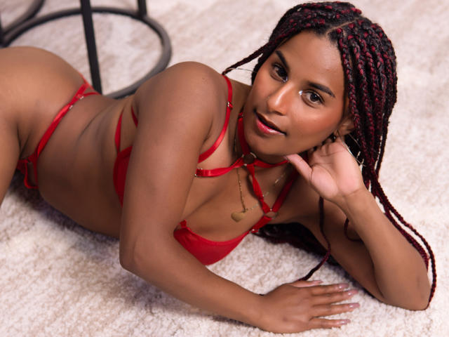 SamanthaRoussee sdraiata su un tappeto, indossa lingerie rossa, ha capelli rossi intrecciati, occhi marroni e una piccola collana d'oro, sorride alla fotocamera, mostrando il suo sedere..