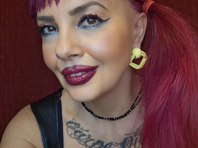 HotAdelayde69 has bright purple hair, blue eyeshadow, thick black eyelashes, shiny purple lipstick, and yellow geometric earrings. Her skin is light and she's smiling.
