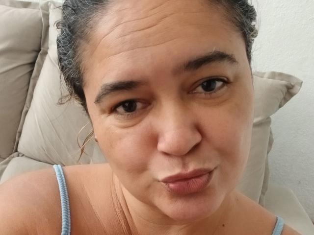 Bethina69 with puffy lips, slightly raised eyebrows, and a cute, playful expression, wearing a blue tank top, has curly dark hair and a light brown skin tone.