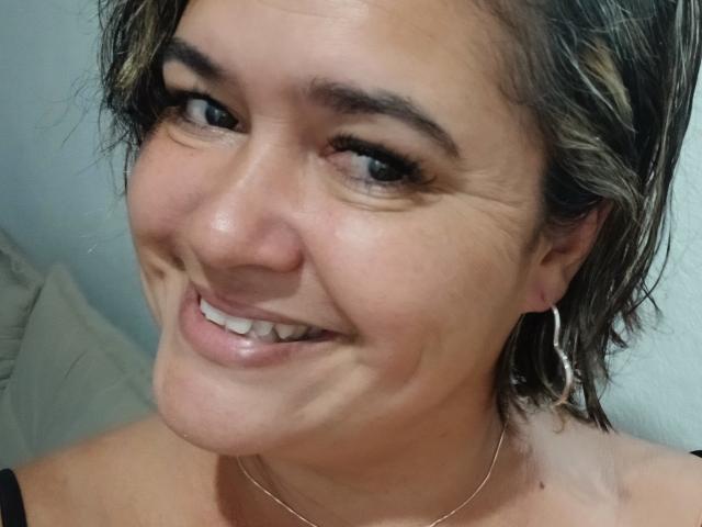 Bethina69 with short brown hair, smiling, showing her teeth, wearing a black top, hoop earrings, and a thin necklace, her ass is not visible. Her face is centered.