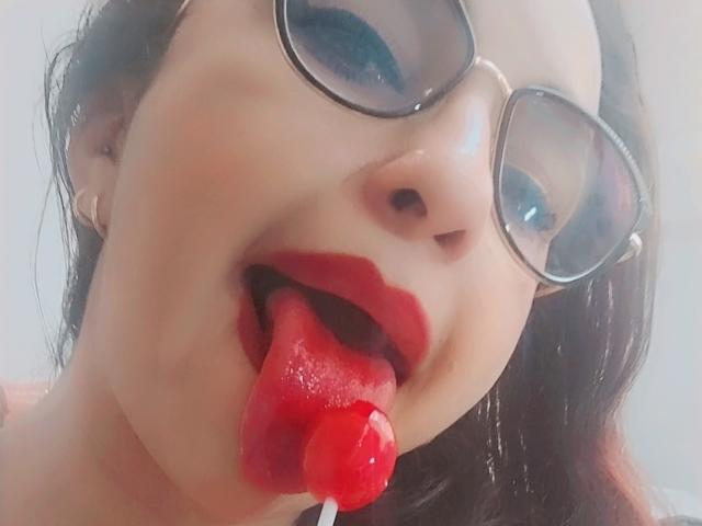 LuluFrancaise with red lips, tongue out, holding a red lollipop, wearing glasses, has fair skin, and dark brown hair, looking up with a playful expression.