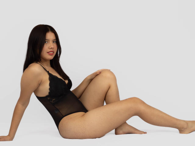 MarielQueenX69 is sitting with her legs spread, wearing a black sheer lingerie that shows her small tits and pussy, against a white background. She has dark brown hair.