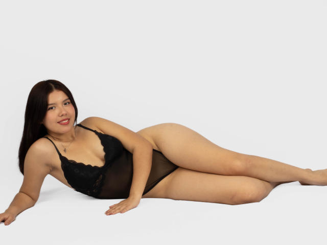 MarielQueenX69 lies on her side in a black sheer lingerie bodysuit, showing her ass, tits, and pussy, with a smile, on a white background.