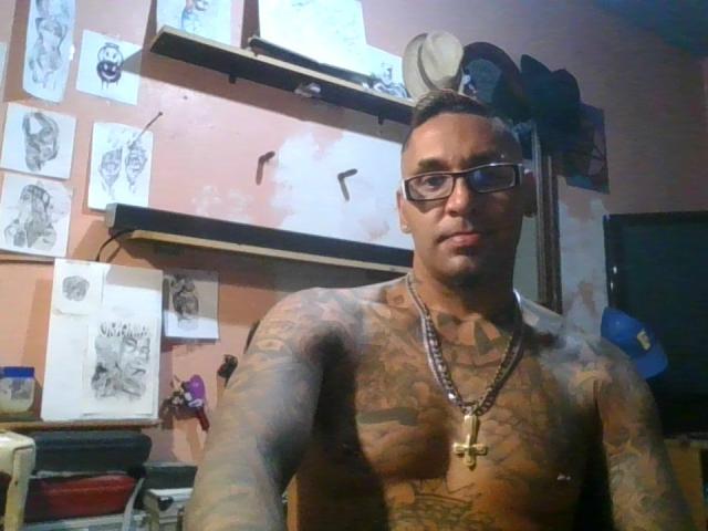 SpunoutAndNaked is a muscular, tattooed dude with glasses, chest tattoo, and cross necklace, standing shirtless in a room with sketch papers and shelves in the background.