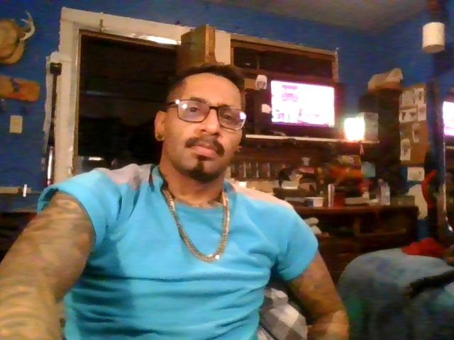 SpunoutAndNaked, a tattooed dude with glasses, mustache, and goatee, wearing a blue shirt and chain, taking a selfie in a dimly lit room.