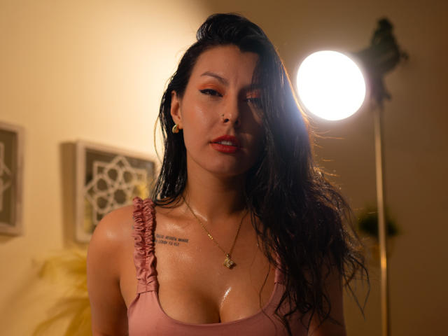 AnnyCraffort with wet black hair, pink tank top, shiny skin, small gold earrings, necklace, red lipstick, and tattoo on right shoulder, standing under bright light, showing her tits.