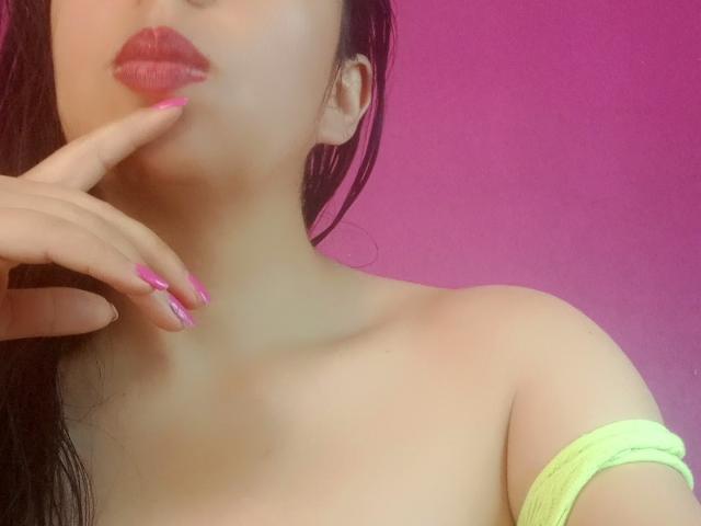 SublimeMature has pink lipstick, finger with matching pink nails touching her lips, neon green strap on her shoulder, and pink background. She has light skin and black wet hair.
