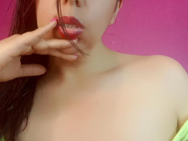 El rostro de SublimeMature con lápiz labial rosa, cubriendo parcialmente su boca con una mano, mostrando sus senos, sobre un fondo rosa, con cabello castaño y una blusa verde con un hombro descubierto..