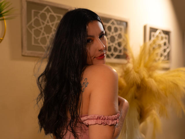 AnnyCraffort with long black hair, pink off-shoulder top, red lipstick, tattoo on right shoulder, looking over shoulder, holding small breast, yellow pampas grass in background.