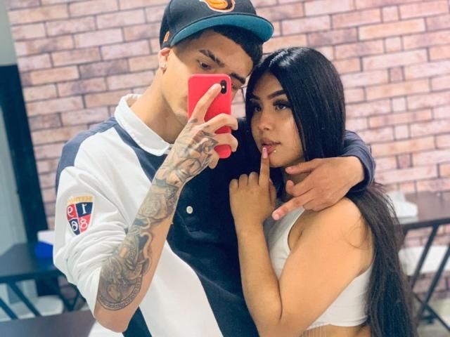 David en Laura, met tatoeages op de linkerarm, met een pet op, een wit shirt aan, en een rode telefoon in de hand, staan naast Laura met lang zwart haar, een witte beha, en een vinger op haar lippen, in een selfie..
