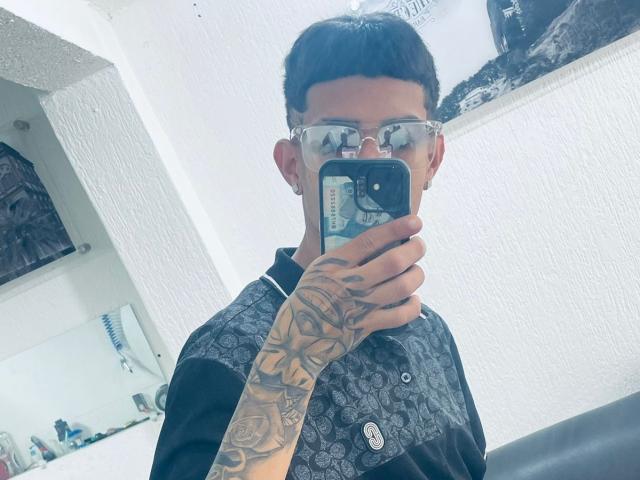 David e Laura, con tatuaggi sul braccio destro, che indossa occhiali, una maglietta nera, tiene il telefono per un selfie allo specchio, parete bianca con texture, piercing all'orecchio nero, capelli neri corti, in una stanza luminosa..