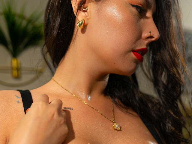 AnnyCraffort's face and upper chest, wet skin with droplets, red lipstick, black hair, gold earrings, nose ring, necklace, "MOTHER" tattoo, right hand on strap, slight profile.