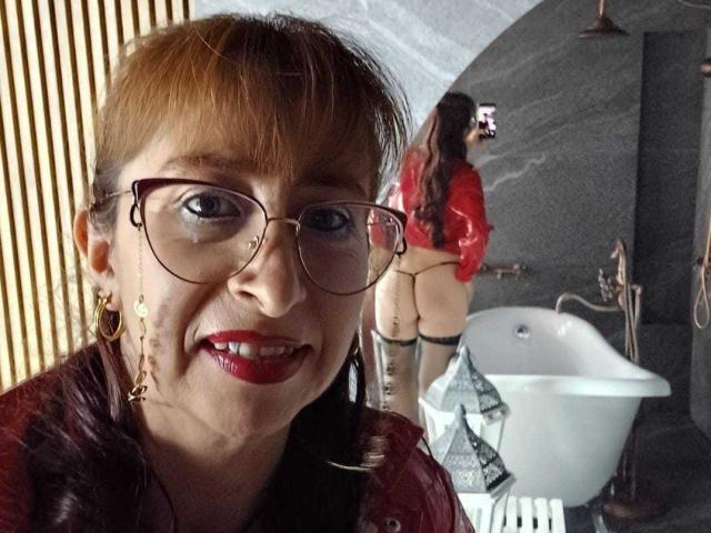 ViktoriaBrown with red hair, glasses, and red lipstick, smiling, wearing a red top, reflected in a mirror showing her bare ass, thigh-high boots, and a red jacket.