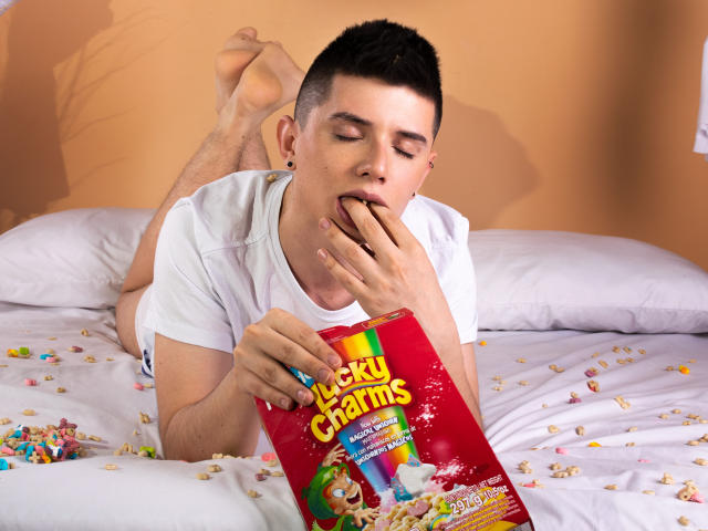 JacobWinds, short dark hair, white t-shirt, eating from a red Lucky Charms box, lying on a bed with scattered cereal, hand in mouth, feet up, relaxed, eating.