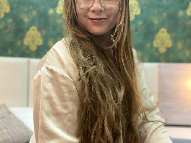 SiaMiller with long brown hair, wearing clear glasses and a shiny pink satin shirt, smiling softly at the camera. Her tits are not visible, just her upper body.