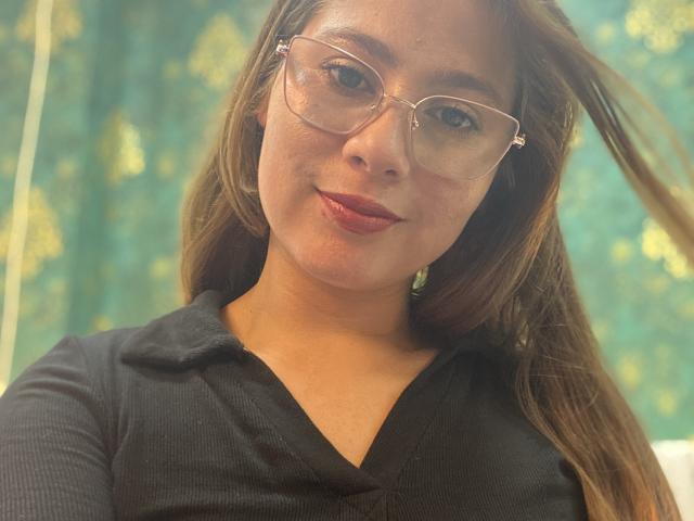 SiaMiller, avec des lunettes, cheveux bruns, chemise noire, un léger sourire, yeux marrons, peau claire, petit nez, maquillage discret, éclairage doux, selfie décontracté..