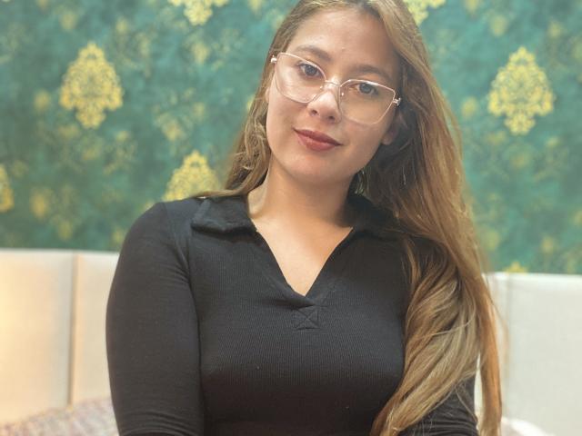 SiaMiller tem cabelos castanhos longos, usa óculos e veste uma camisa preta com decote em V, que mostra seus seios, com um leve sorriso, sentada em um cômodo bem iluminado..