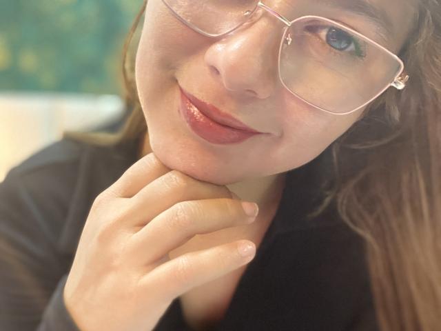 SiaMiller with glasses, light brown hair, and pink lipstick, holding her chin with a cute smile, wearing a black shirt, showing her tits slightly in the frame.