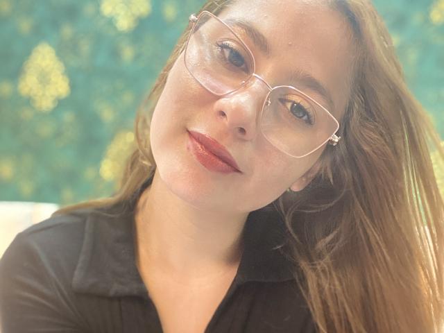 SiaMiller with glasses, light brown hair, and red lipstick, wearing a black shirt, smiling softly at the camera, sunlight highlighting her face.