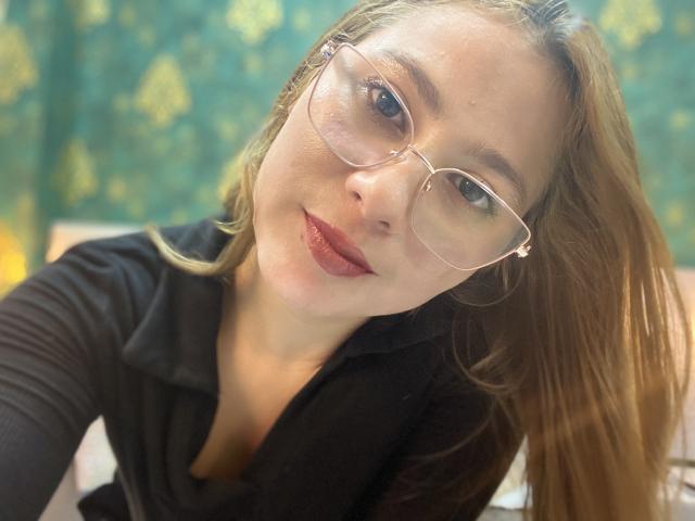SiaMiller con gafas, cabello castaño claro, lápiz labial rojo, camisa negra, inclinada hacia adelante, sonriendo ligeramente, luz suave, escote visible..