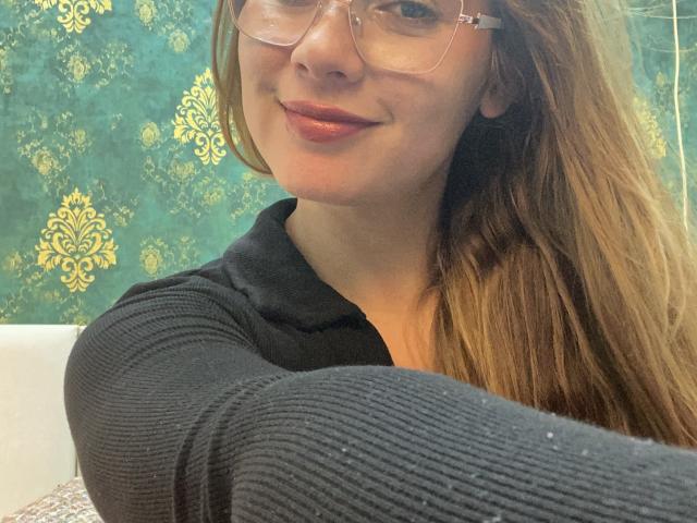 SiaMiller with glasses, long brown hair, smiling, wearing a black ribbed shirt, showing her tits in a selfie, red lipstick, and a cute, attractive face.