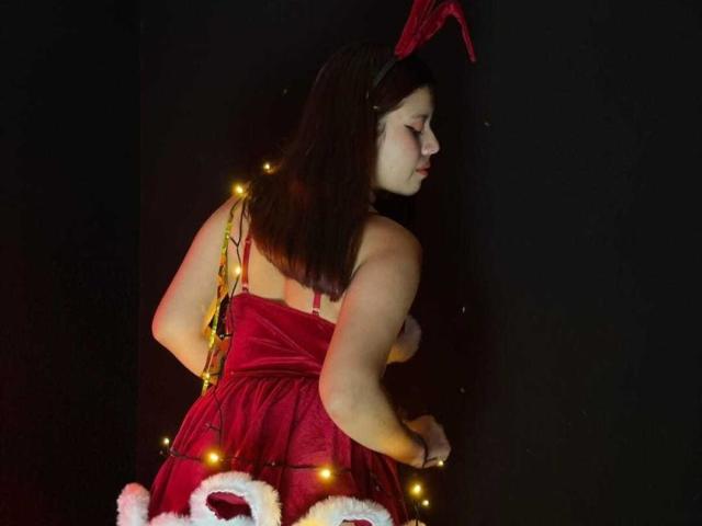 MarielQueenX69 with red velvet dress, red bunny ears, and Christmas lights, her ass slightly visible, dark hair, facing black wall, soft light, sexy, curvy, looking away.
